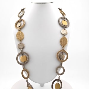 May include: A long necklace with a mix of gold and silver tones. The necklace features a series of circular and oval-shaped pendants, some with textured patterns. The necklace is displayed on a white mannequin, showcasing its design and length.