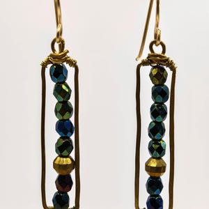 May include: A pair of handmade earrings with a rectangular design. Each earring features a series of faceted, iridescent beads in shades of blue, green, and gold, strung within a gold-toned wire frame. The earrings hang from gold-colored ear wires.