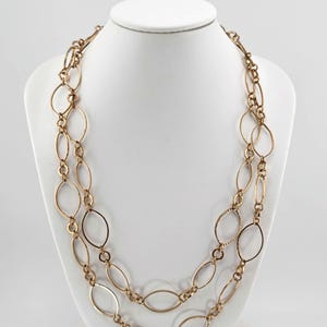 May include: A gold-toned necklace with two strands of oval-shaped links. The necklace is displayed on a white bust form against a white background. The links have a textured design.
