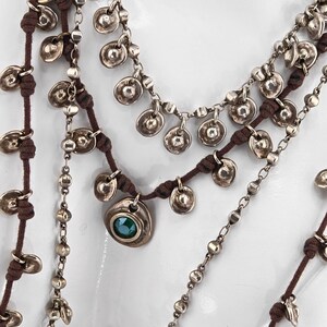 May include: Multiple layered necklaces with silver-toned metal discs and beads. The necklaces are strung on brown cords. One necklace features a pendant with a green stone. The jewelry is displayed against a white background.