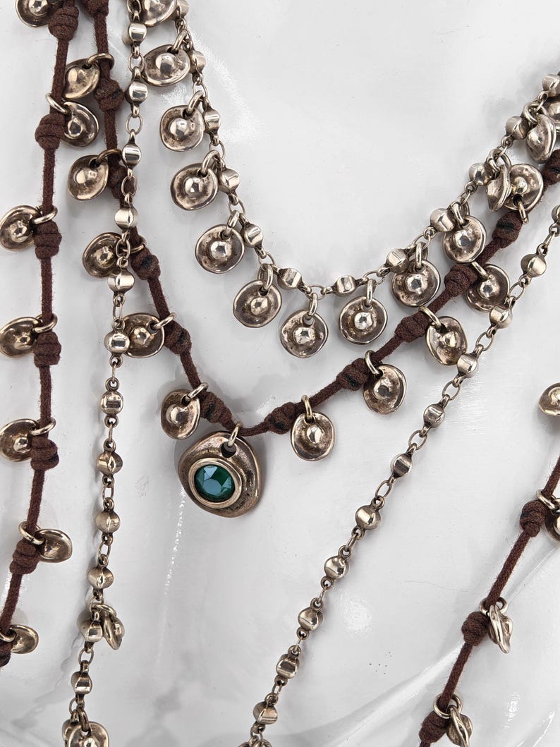 May include: Multiple layered necklaces with silver-toned metal discs and beads. The necklaces are strung on brown cords. One necklace features a pendant with a green stone. The jewelry is displayed against a white background.
