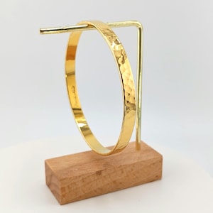 May include: A gold-colored bangle bracelet with a hammered texture is displayed on a wooden stand. The bracelet has a smooth, polished interior and a textured exterior. The stand is made of a light-colored wood and has a gold-colored metal support.