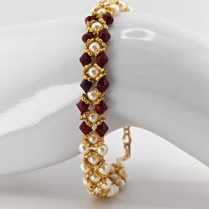 May include: A gold-toned bracelet featuring alternating rows of white pearls and deep red, faceted beads. The bracelet is accented with gold-colored filigree and has a delicate chain clasp. The jewelry is displayed on a white surface.