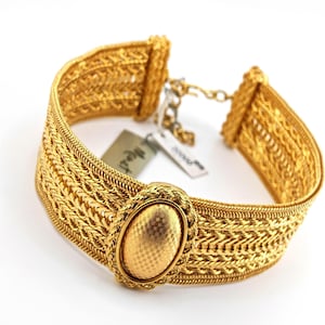 May include: A gold-tone metal choker necklace featuring a wide, woven mesh band and a central oval medallion with a textured, cross-hatched surface. The piece includes a lobster claw clasp, an adjustable chain, and two small rectangular tags labeled Monet.