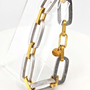 May include: A silver and gold-toned chain link bracelet. The bracelet features rectangular links in silver and gold, connected by smaller gold links. A small, round gold charm hangs from the clasp.