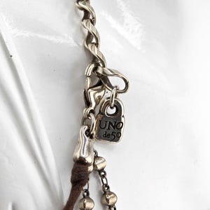 May include: Close-up of a silver-toned necklace with a twisted chain design. The necklace features a small rectangular charm with the text "UNO de 50". A brown cord and a chain with small, round beads are also visible.