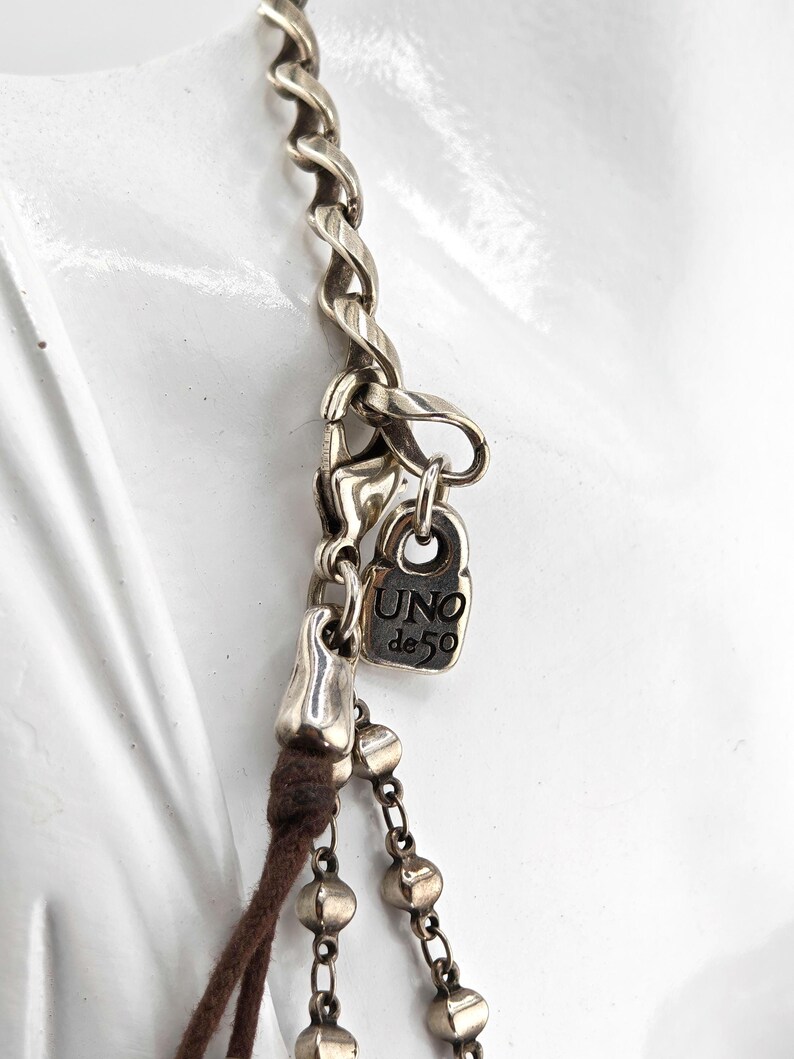 May include: Close-up of a silver-toned necklace with a twisted chain design. The necklace features a small rectangular charm with the text "UNO de 50". A brown cord and a chain with small, round beads are also visible.