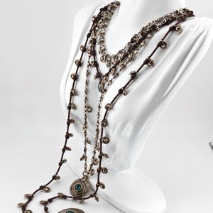 May include: Multiple layered necklaces with silver-toned pendants and beads. The necklaces are displayed on a white mannequin. The pendants have a circular design with a teal-colored stone in the center. The necklaces have brown cords.