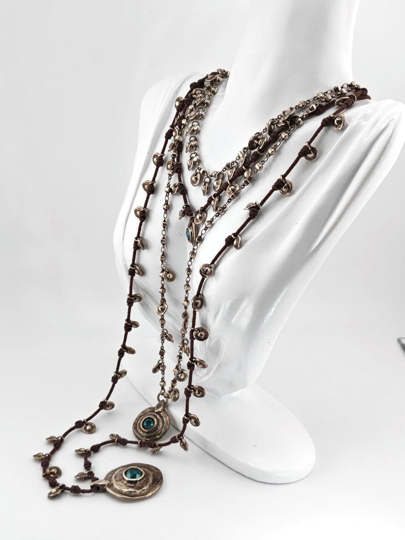 May include: Multiple layered necklaces with silver-toned pendants and beads. The necklaces are displayed on a white mannequin. The pendants have a circular design with a teal-colored stone in the center. The necklaces have brown cords.