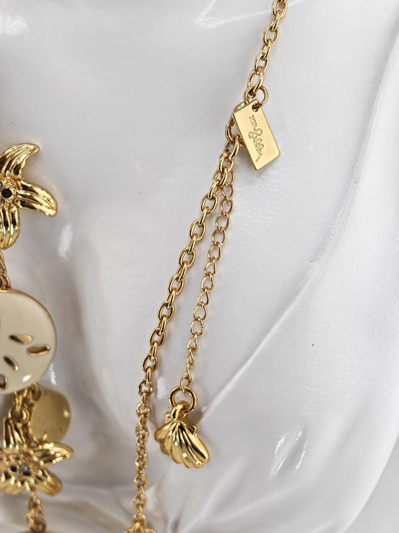 May include: Gold-toned necklace with seashell and starfish charms. The necklace features a delicate chain and a small tag with the brand name. The charms are detailed and add a coastal theme to the jewelry.