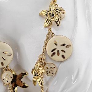 May include: Gold-toned necklace featuring starfish and sand dollar charms. The starfish charms have black accents and the sand dollar charms are cream-colored. The necklace is designed with a delicate chain and is suitable for beach-themed jewelry.