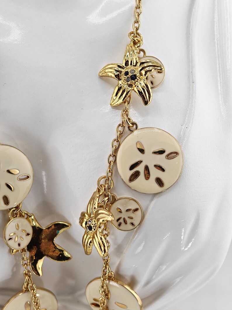 May include: Gold-toned necklace featuring starfish and sand dollar charms. The starfish charms have black accents and the sand dollar charms are cream-colored. The necklace is designed with a delicate chain and is suitable for beach-themed jewelry.