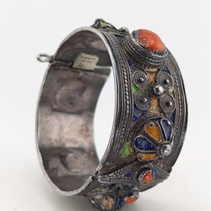 May include: A silver-toned cuff bracelet with intricate enamel and gemstone details. The bracelet features a central design with orange, blue, green, and yellow enamel, and is accented with small red gemstones. The bracelet has a hinged closure.