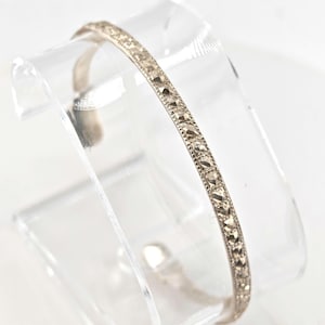 May include: A silver-toned bracelet with a repeating geometric pattern of small, square-shaped embellishments. The bracelet is displayed on a clear, curved stand, showcasing its delicate design and craftsmanship. The bracelet appears to be made of metal.
