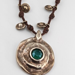 May include: A bronze-toned necklace with a large, textured pendant featuring a central, round, emerald-green gemstone. The pendant is suspended from a brown cord, adorned with small, bronze-colored disc charms.