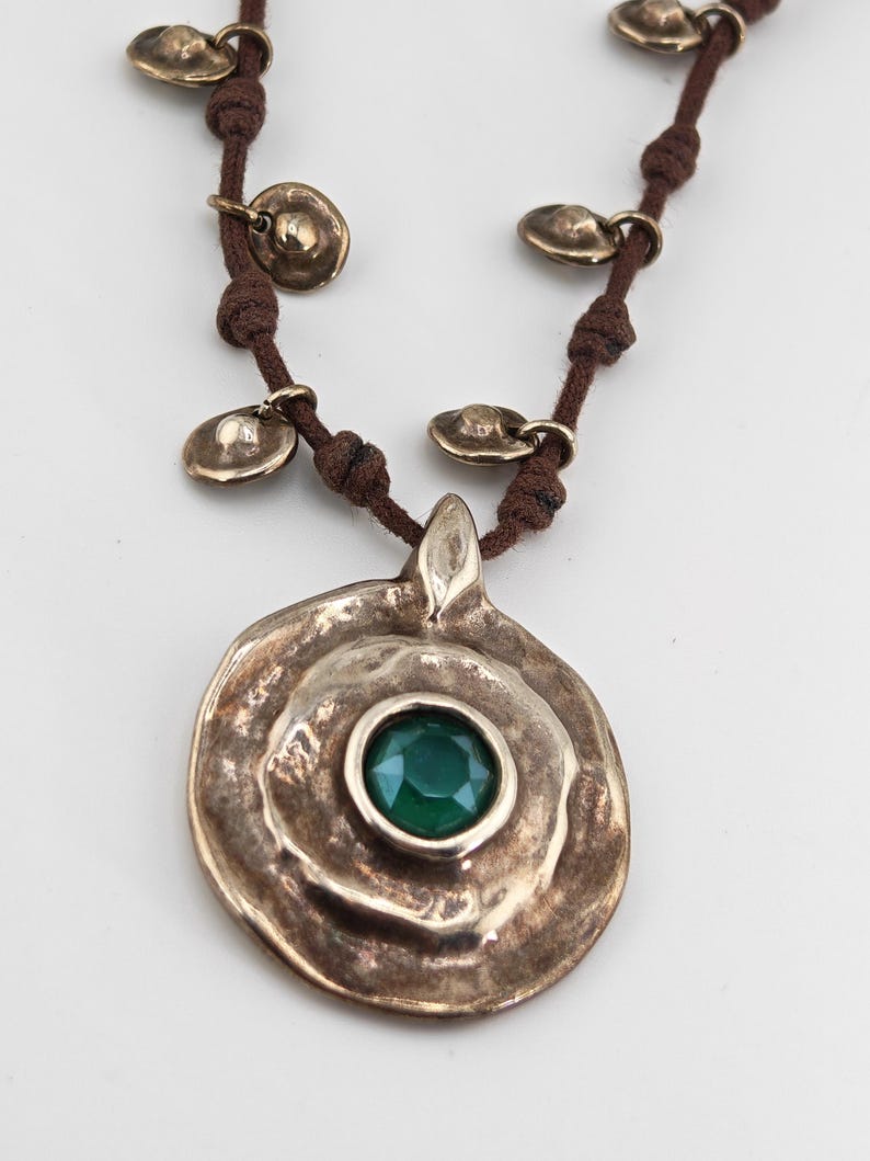 May include: A bronze-toned necklace with a large, textured pendant featuring a central, round, emerald-green gemstone. The pendant is suspended from a brown cord, adorned with small, bronze-colored disc charms.