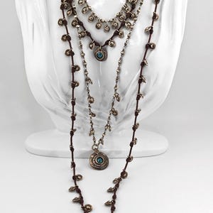 May include: Three layered necklaces with a bohemian style. The necklaces are displayed on a white bust. The necklaces have a brown cord and silver-colored metal pendants. Each necklace has a different design, with turquoise-colored stones.