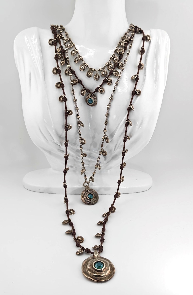 May include: Three layered necklaces with a bohemian style. The necklaces are displayed on a white bust. The necklaces have a brown cord and silver-colored metal pendants. Each necklace has a different design, with turquoise-colored stones.