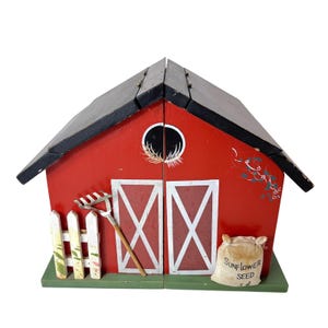 May include: A red wooden barn-shaped decorative box with a black roof. The front features white double doors, a white picket fence, a metal rake, and a small sack labeled Sunflower Seed 5 cents. A circular window is painted near the roof peak.