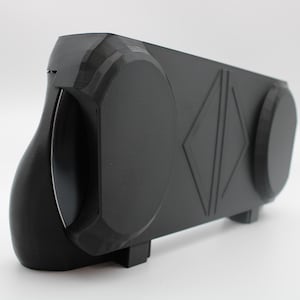 Puede incluir: Un soporte rectangular de plástico negro mate con dos montajes laterales circulares y un detalle central en relieve en forma de diamante. El objeto tiene un lado izquierdo curvo y una base plana con pequeños pies de apoyo.