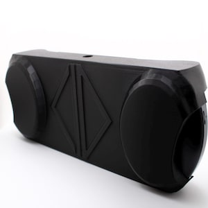 May include: A black, rectangular object with rounded ends and a geometric design. The object has a diamond-shaped accent in the center and a small hole on top. It is placed on a white surface.