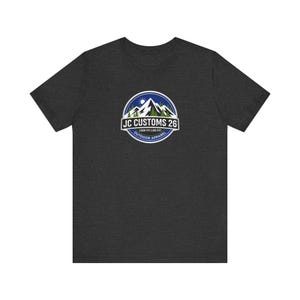 Puede incluir: Camiseta gris oscuro con un logotipo circular que representa montañas, un sol y texto. El logotipo dice "JC CUSTOMS 26", "LOOK FIT LIVE FIT" y "OUTDOOR APPAREL". La camiseta es de cuello redondo y manga corta.