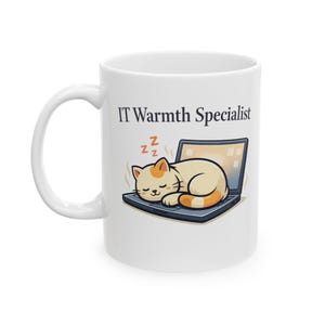 Puede incluir: Taza de cerámica blanca con el texto "IT Warmth Specialist" y una ilustración de dibujos animados de un gato durmiendo en un portátil. El gato es de color crema con marcas naranjas. La pantalla del portátil está encendida.