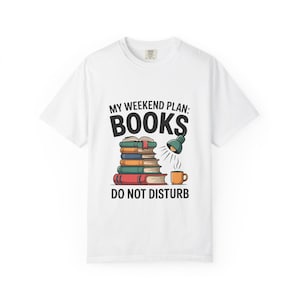 Op de afbeelding: Wit t-shirt met een grafisch ontwerp. Het ontwerp toont een stapel boeken, een groene bureaulamp en een koffiekopje. De tekst op het shirt luidt "MY WEEKEND PLAN: BOOKS DO NOT DISTURB."