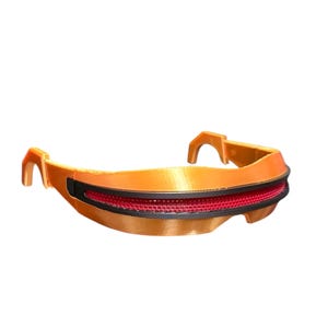 May include: A visor-style eyewear piece featuring a metallic orange frame with a smooth, curved design. The center section contains a horizontal black trim piece that houses a textured, vibrant red mesh lens spanning across the front of the frame.