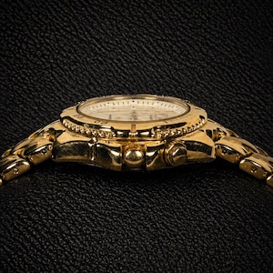 May include: A gold-toned wristwatch with a round face and a linked band. The watch face is white with black markings and hands. The bezel is adorned with small, decorative elements. The watch is set against a dark, textured background.
