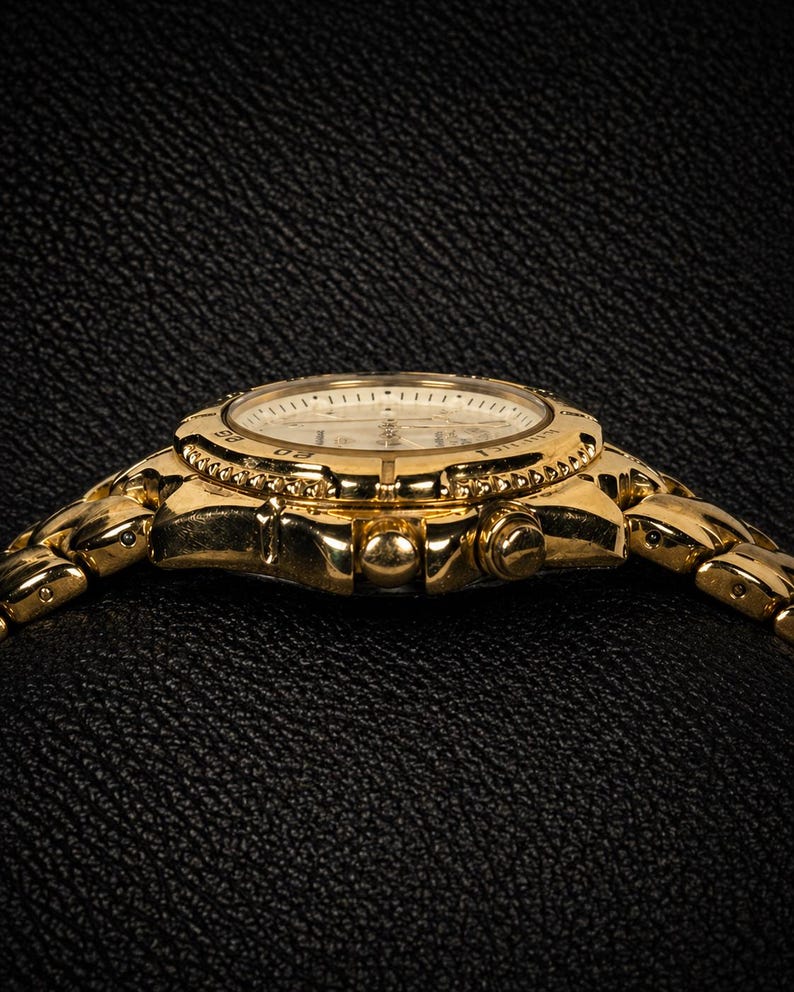 May include: A gold-toned wristwatch with a round face and a linked band. The watch face is white with black markings and hands. The bezel is adorned with small, decorative elements. The watch is set against a dark, textured background.