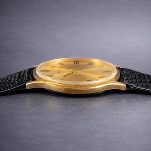 May include: A gold-toned wristwatch with a black band. The watch face has Roman numerals and gold hands. The watch band is made of a textured black material. The watch is a classic design.