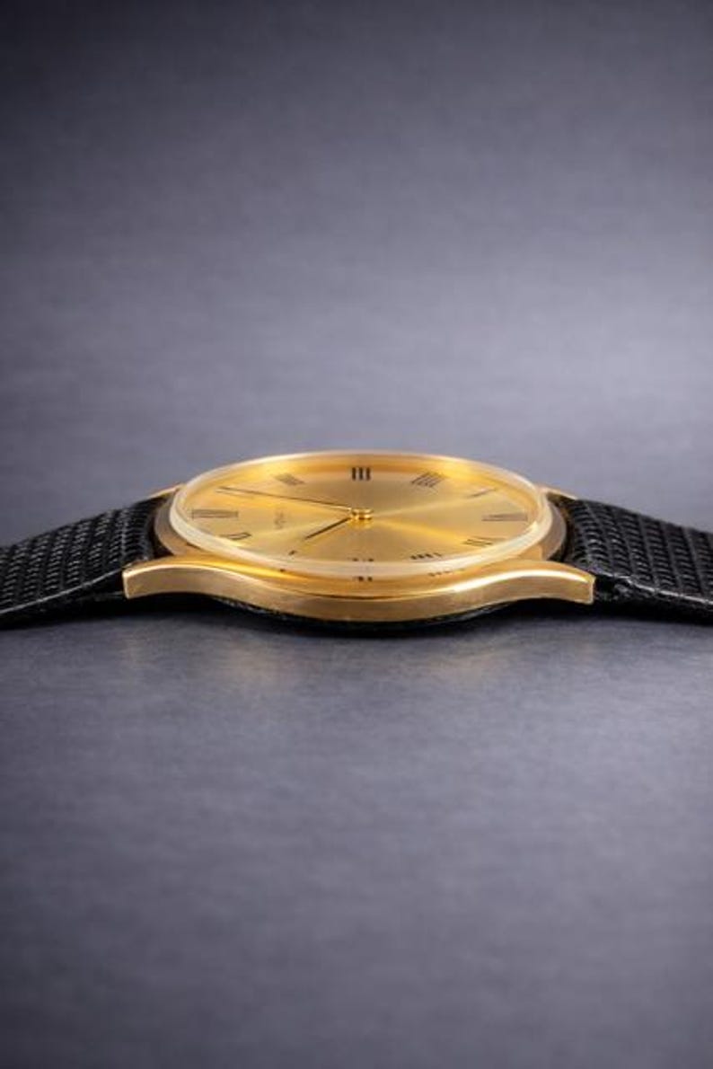 May include: A gold-toned wristwatch with a black band. The watch face has Roman numerals and gold hands. The watch band is made of a textured black material. The watch is a classic design.