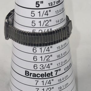 May include: A silver-toned watch bracelet is placed on a white bracelet sizing cone. The cone displays measurements from 5" (12.7 cm) to 8 3/4" (22.2 cm), with both inches and centimeters. The text "Bracelet 7" is visible.