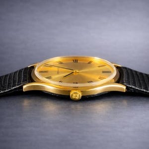 May include: A gold-toned wristwatch with a black leather band. The watch face has Roman numerals and a minimalist design. The watch case is gold, and the band is textured. The watch is a classic accessory.