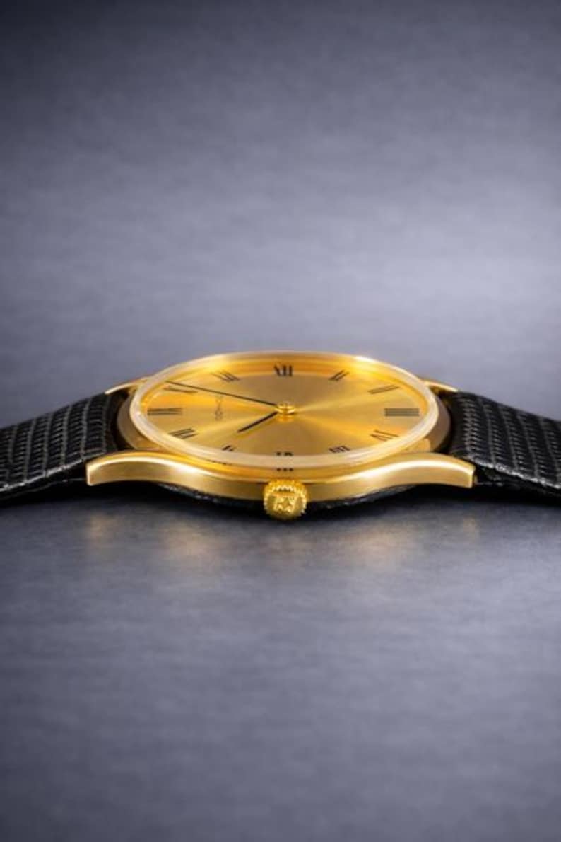 May include: A gold-toned wristwatch with a black leather band. The watch face has Roman numerals and a minimalist design. The watch case is gold, and the band is textured. The watch is a classic accessory.