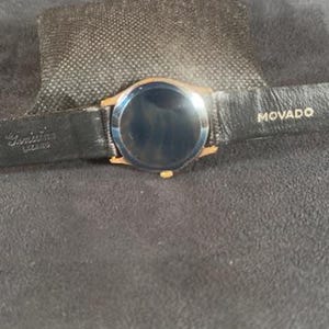 May include: A gold-toned Movado watch with a black leather band. The watch face is dark, and the band is marked with the Movado brand name. The watch is set on a dark gray surface.