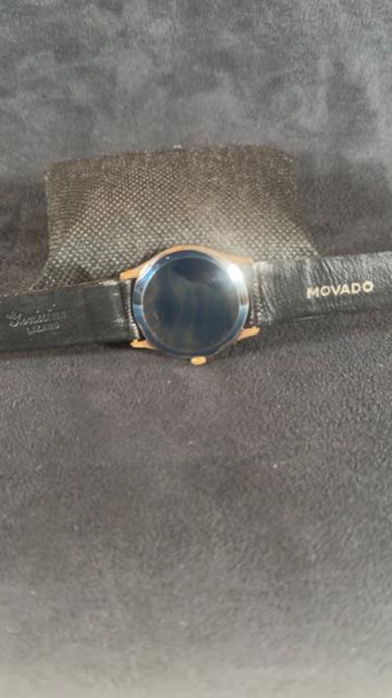 May include: A gold-toned Movado watch with a black leather band. The watch face is dark, and the band is marked with the Movado brand name. The watch is set on a dark gray surface.