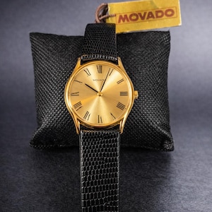 May include: A gold-toned Movado watch with a black leather band. The watch face has Roman numerals and a minimalist design. The watch is displayed on a black textured pillow, with a Movado tag.