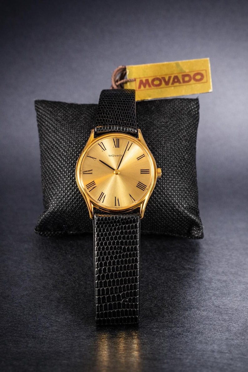 May include: A gold-toned Movado watch with a black leather band. The watch face has Roman numerals and a minimalist design. The watch is displayed on a black textured pillow, with a Movado tag.