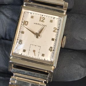 May include: A vintage Hamilton rectangular wristwatch with a gold-tone case and a matching flexible metal expansion band. The off-white dial features gold-tone Arabic numerals at the 12, 10, 8, 4, and 2 positions, with a small sub-dial for seconds.