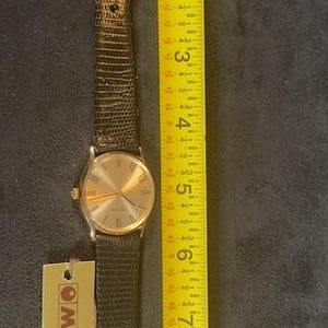 May include: A gold-toned Movado watch with a black leather band, next to a yellow measuring tape. The watch face has gold hands and Roman numerals. The watch band is approximately 8 inches long.