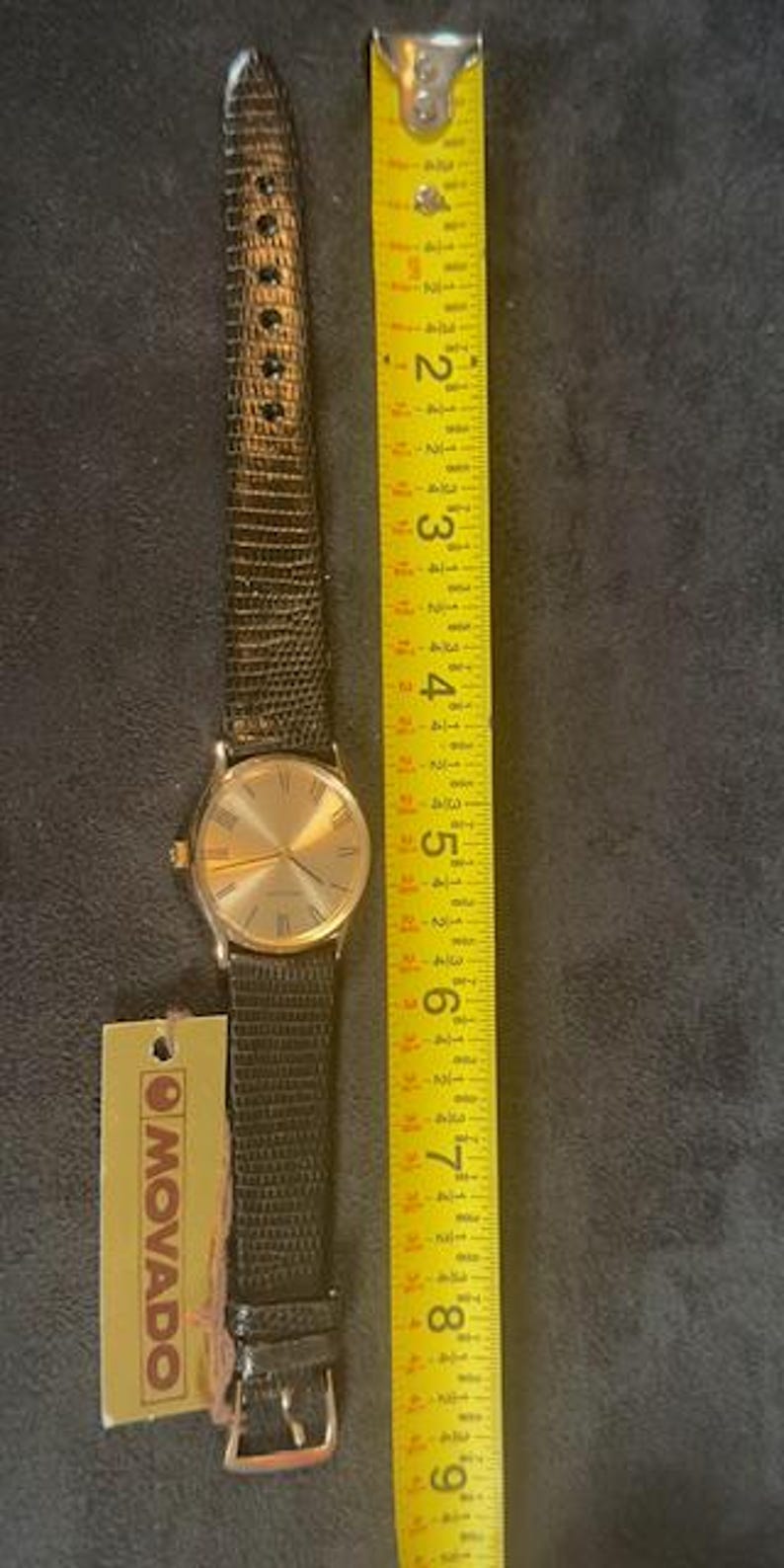 May include: A gold-toned Movado watch with a black leather band, next to a yellow measuring tape. The watch face has gold hands and Roman numerals. The watch band is approximately 8 inches long.
