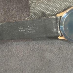 May include: Close-up of a vintage wristwatch with a black leather band. The band is stamped with the words "Genuine Lizard". The watch face is a dark color with gold-toned accents. The watch is set against a dark gray background.