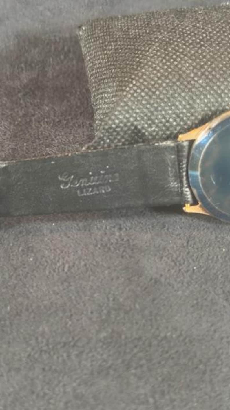 May include: Close-up of a vintage wristwatch with a black leather band. The band is stamped with the words "Genuine Lizard". The watch face is a dark color with gold-toned accents. The watch is set against a dark gray background.