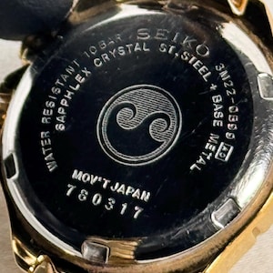 May include: Close-up of a gold-toned Seiko watch. The watch back is black with white text, including "Water Resistant," "Sapphlex Crystal," and "Mov't Japan." A stylized yin-yang symbol is in the center. The watch band is also gold.