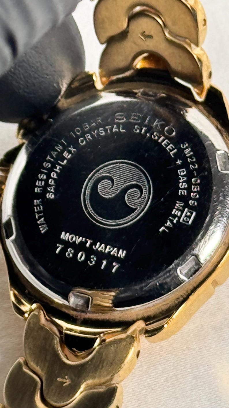 May include: Close-up of a gold-toned Seiko watch. The watch back is black with white text, including "Water Resistant," "Sapphlex Crystal," and "Mov't Japan." A stylized yin-yang symbol is in the center. The watch band is also gold.