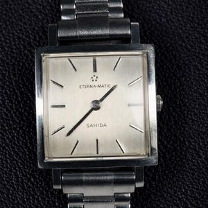May include: A vintage Eterna-Matic Sahida wristwatch with a silver square face and a stainless steel band. The watch face features black hands and markers. The watch is displayed on a dark textured surface.