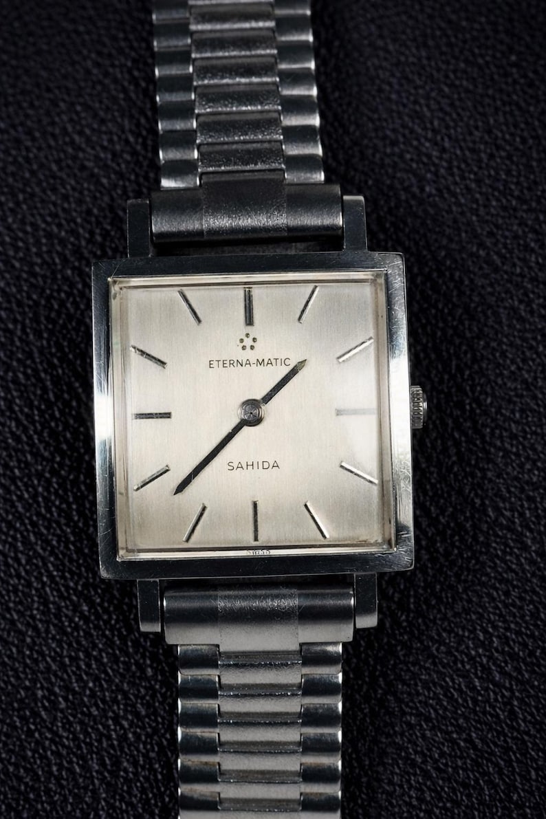 May include: A vintage Eterna-Matic Sahida wristwatch with a silver square face and a stainless steel band. The watch face features black hands and markers. The watch is displayed on a dark textured surface.