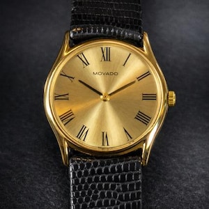 May include: Gold-toned Movado watch with a black leather band. The watch face features Roman numerals and the Movado logo. The band has a textured, reptilian pattern. The watch has a classic, elegant design.